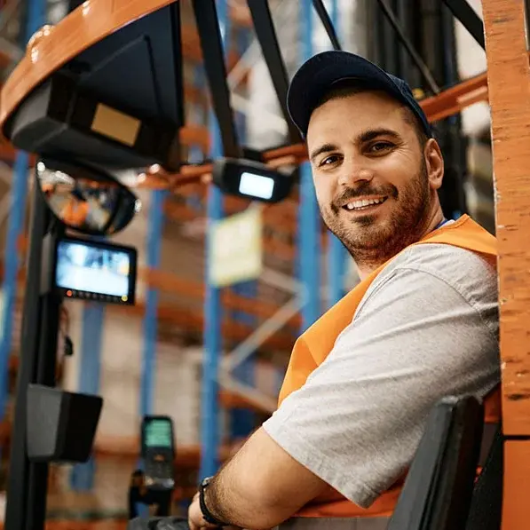 Ein lächelnder Mann mit Bart und Baseballkappe steht in einem Lagerhaus und trägt eine orangefarbene Sicherheitsweste. Er ist wahrscheinlich ein Mitarbeiter von Hermes Fulfilment, der für einen reibungslosen Logistikbetrieb sorgt.